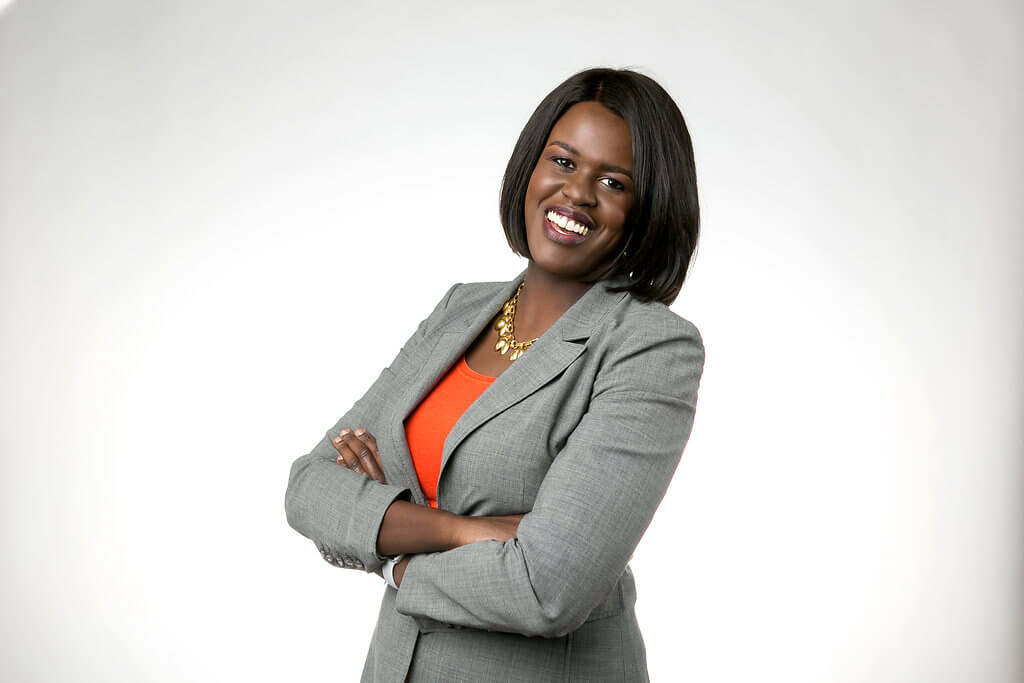 A headshot of Atima Omara standing with her arms crossed. She wears a gray suit jacket with an orange shirt and a gold flower necklace.