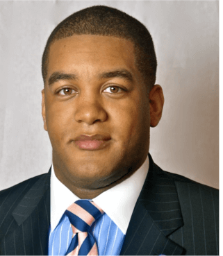 Khalil Thompson, standing 6 feet, 2 inches, with brown hair and oddly blondish beard for a black man. Wearing a blue shirt and white shirt in a suit and tie.