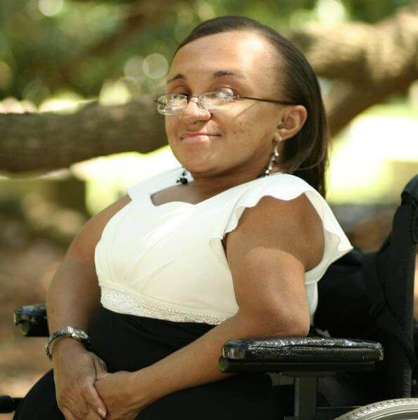 Photo of Vilissa Thompson, a young Black woman with her hair parted and hanging down straight. She is smiling and facing the camera sideways in her wheelchair. She is outside under the tree branches while wearing a white and black dress.