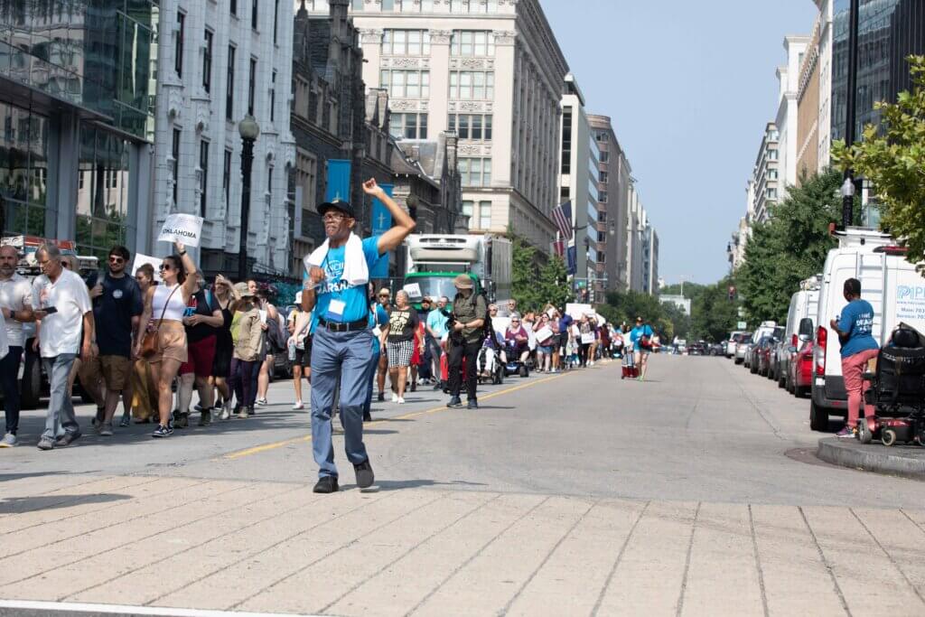 In the background, hundreds of people line the streets of Washington, DC as they march to the U.S. Capitol. In the foreground, NCIL Board Member John Herring raises his fist as he leads marchers as a NCIL Marshall.