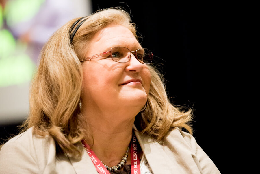 A white person with shoulder length blonde hair and glasses smiles while looking up and away from the camera.