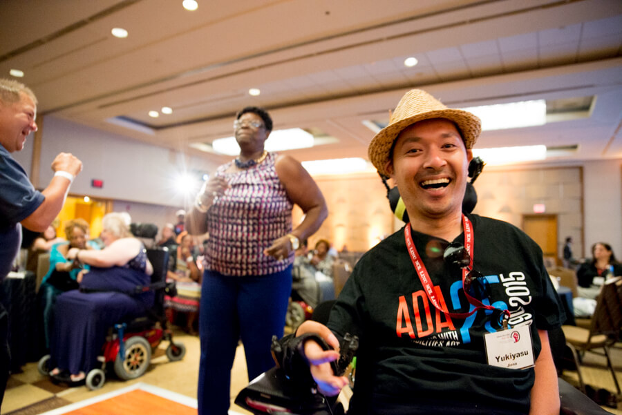 A japanese advocate wearing a straw hat, using a power wheelchair and wearing a nametag that says "Yukiyasu" smiles widely at the camera.