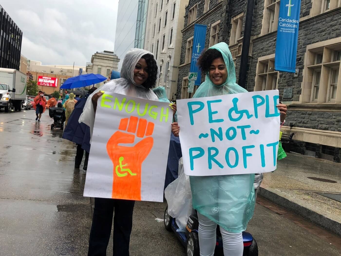 Advocates marching in the rain hold signs that say "Enough" with a raised fist with the international symbol of accessibility and "People not profit" with the O replaced with the international symbol of accessibility.