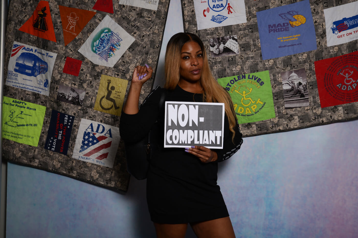 A Black advocate with brownish-blonde hair past their shoulders holds a sign in front of the ADA quilt that says "non-compliant".