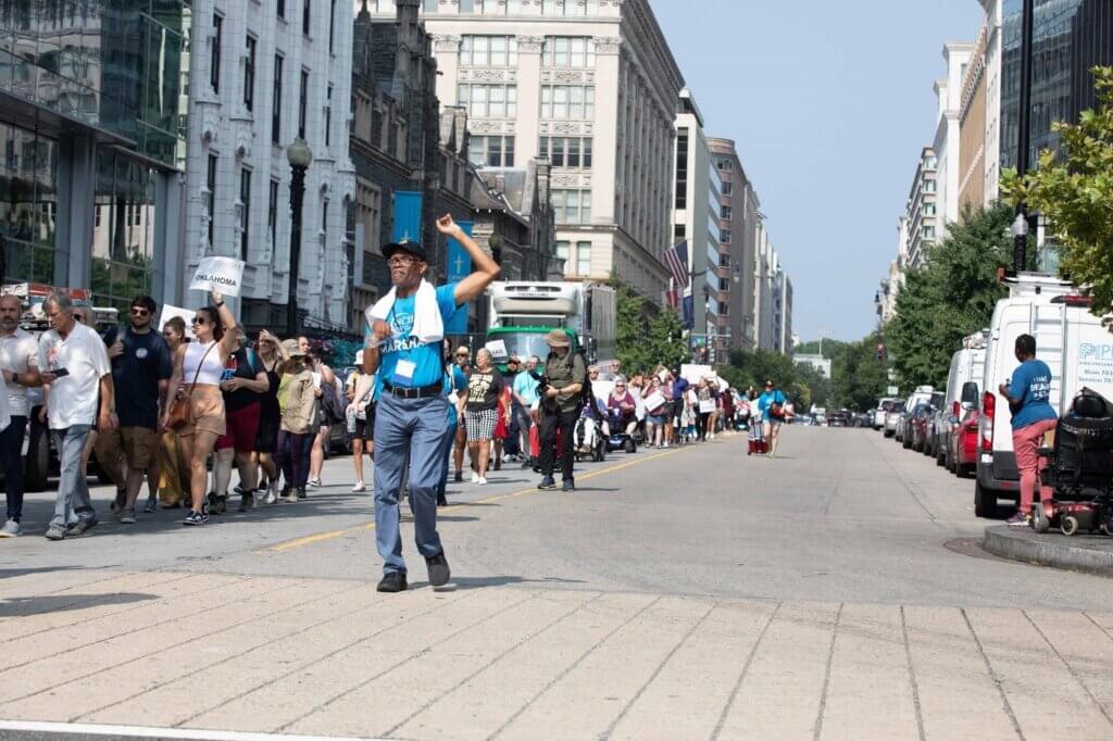 Hundreds of people march to the Capitol. In the foreground, NCIL Marshall John Herring raises his fist as he leads marchers.