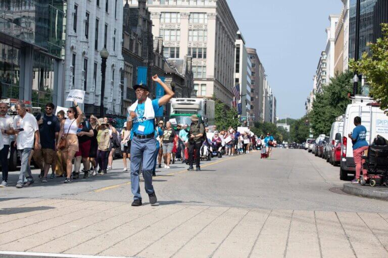 Hundreds of people march to the Capitol. In the foreground, NCIL Marshall John Herring raises his fist as he leads marchers.