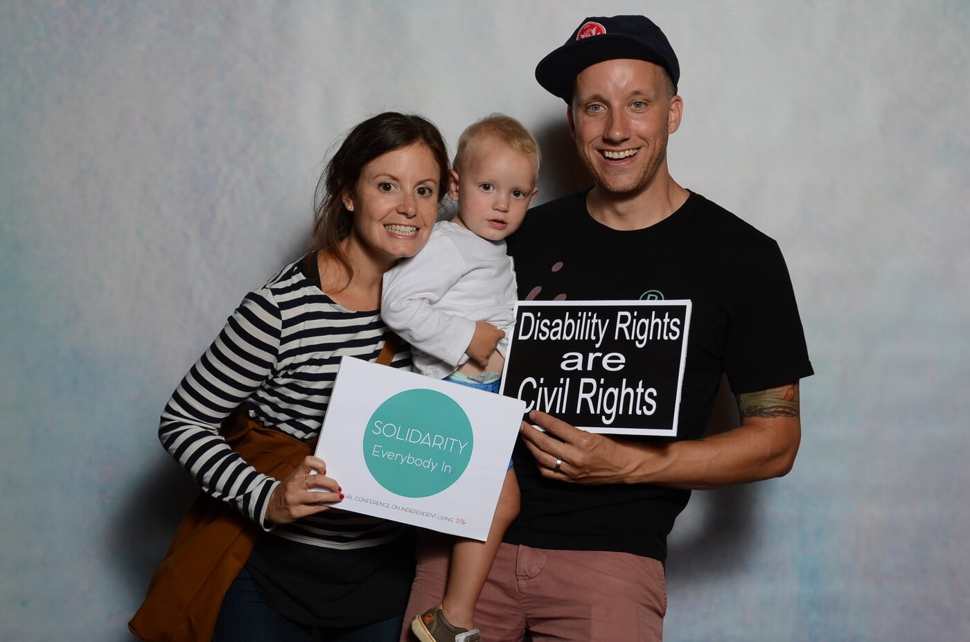 Tim Fuchs, wife Alice, and son Emmett pose at the Annual Conference on Independent Living with signs that say "Solidarity Everybody In" and "Disability Rights Are Civil Rights".