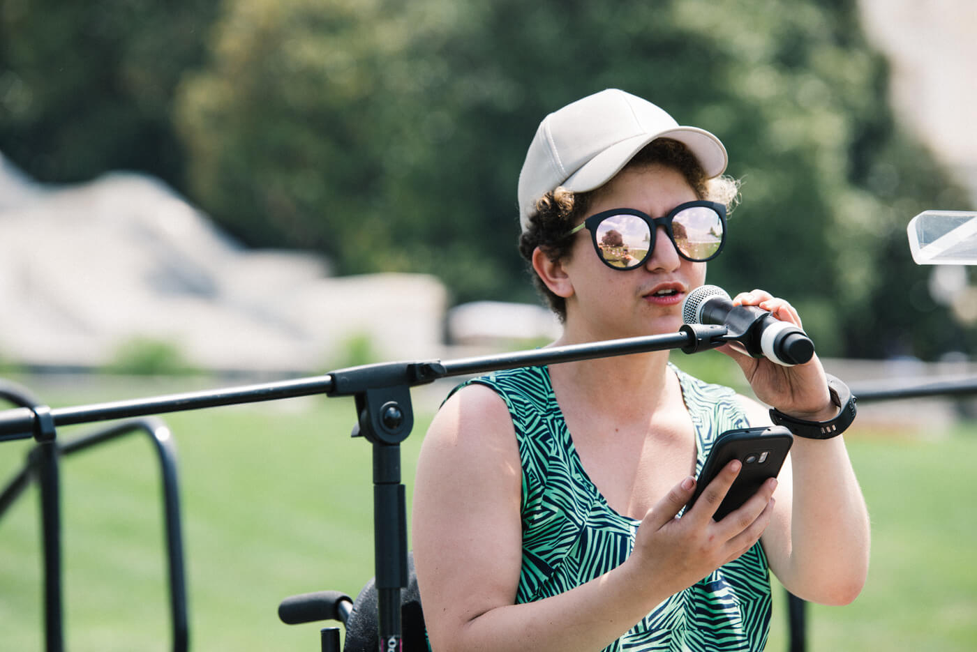 An advocate with short curly brown hair and a white ball cap looks at their phone and speaks into a microphone during NCIL's annual rally.