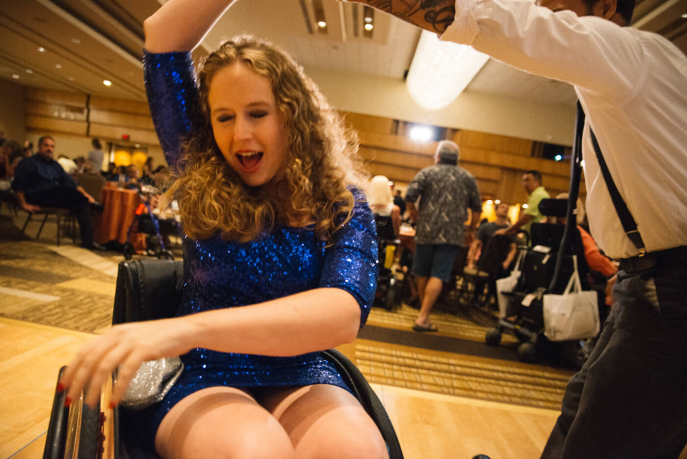 Stephanie Woodward, a white woman with curly blonde hair wearing a sparkly blue dress using a manual wheelchair, dances at the NCIL closing ceremony.
