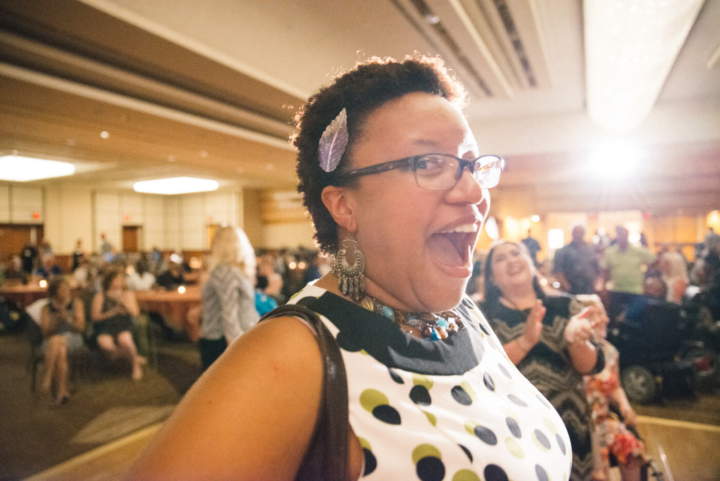 A Black person with short black hair and glasses smiles open mouthed at the camera.