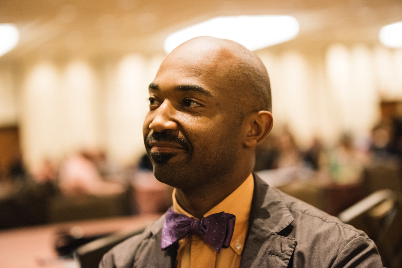A Black person wearing a yellow polo shirt and a polka dot purple bow tie looks off to the side.