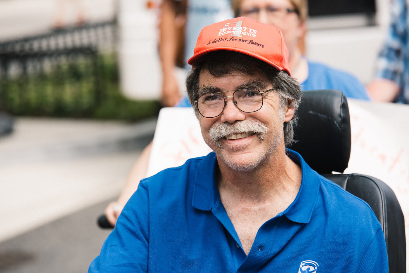 A person with short grey hair, glasses, and a grey mustache wearing an orange "Invest in independence, a dollar for our future" hat and a blue polo shirt using a power wheelchair smiles at the camera.