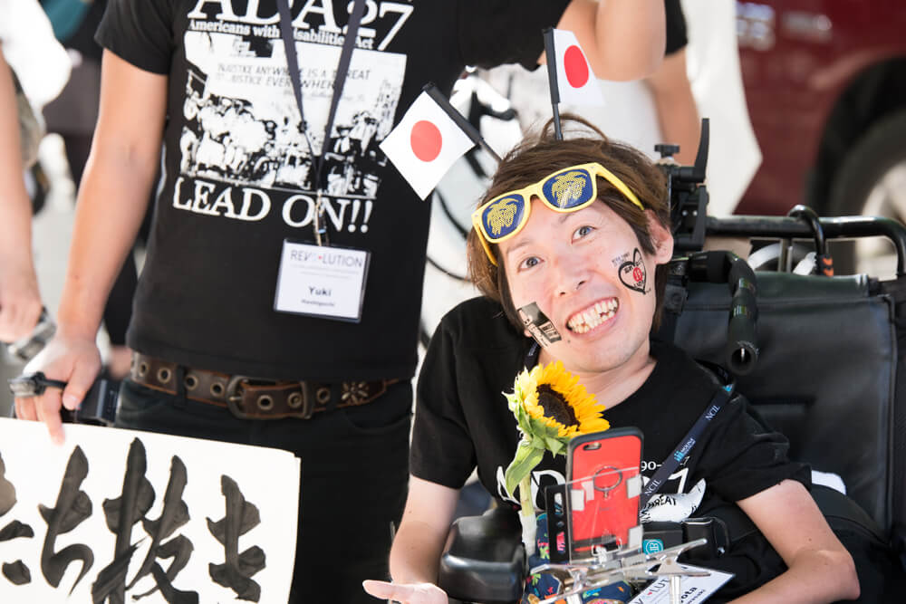 A Japanese advocate using a power wheelchair smiles widely at the camera.