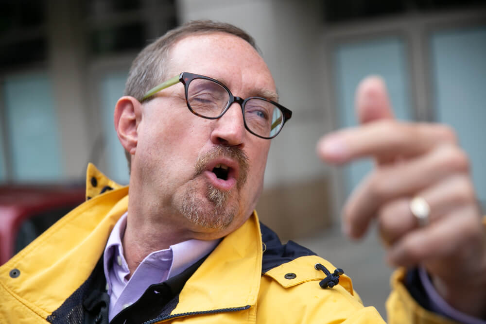 A person with thinning brownish grey hair and glasses wearing a yellow jacket shouts during NCIL's march.