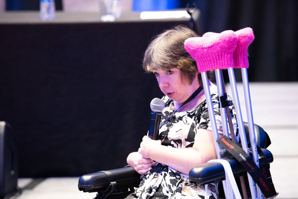 Julie Espinoza speaks into a microphone. Underarm crutches with pink fuzzy tops are visible next to her in her wheelchair.