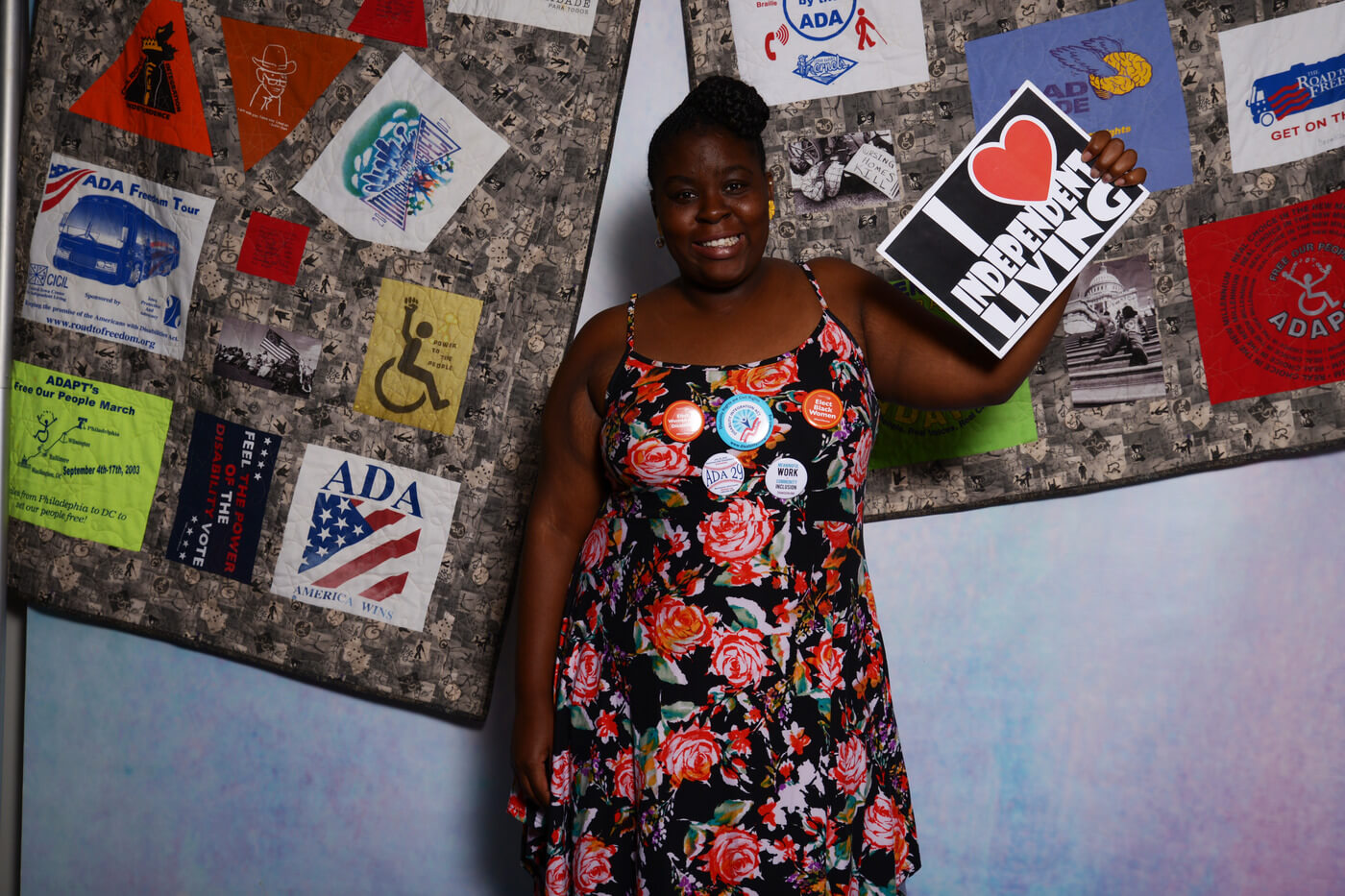 Denise Law stands in front of the ADA quilt and holds a sign that says "I [heart] Independent Living".