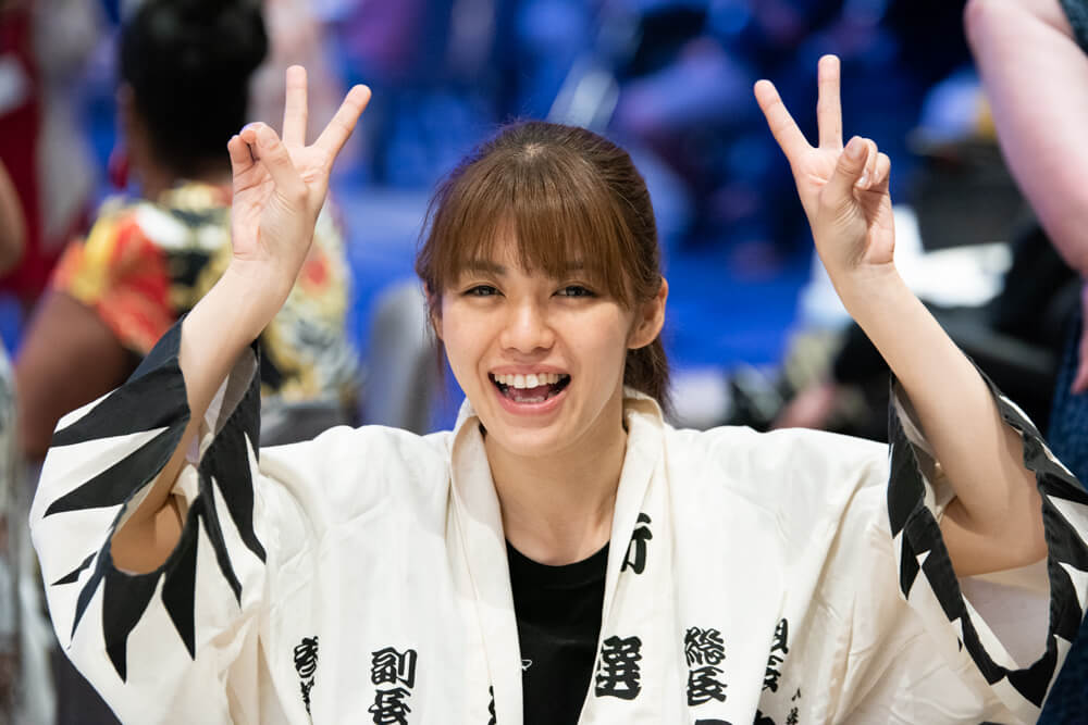 A Japanese advocate with brown hair pulled back into a ponytail shows double peace signs to the camera.