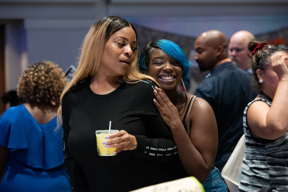 Two young Black women, one holding a drink and one slightly behind the other, pose for a picture.