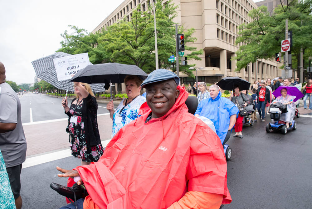 An advocate wearing a red rain poncho smiles at the camera. In the background, other advocates march and one holds a sign that says "North Carolina".