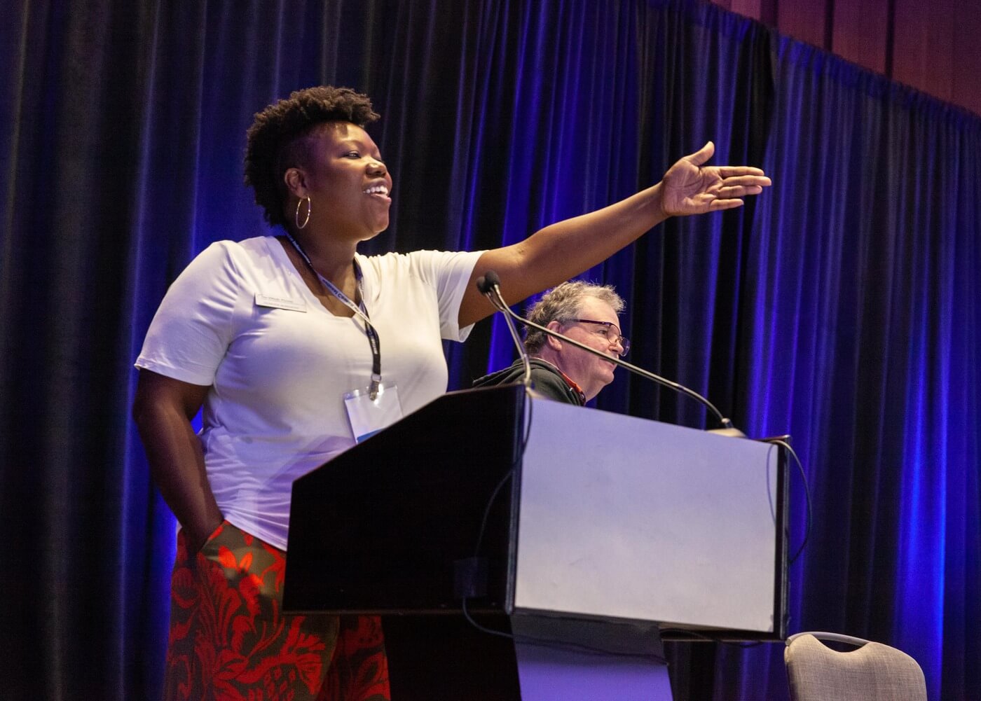 A Black person with short, brown, curly hair and large hoop earrings stands at a podium next to Steve Higgins and gestures out at the audience.