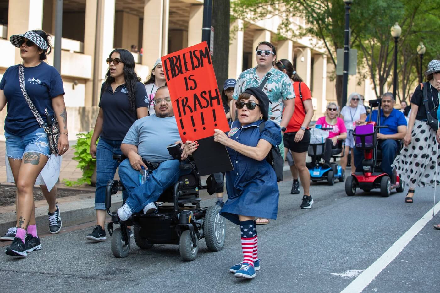 A white Little Person advocate marches with a sign that says "Ableism is Trash!!!"