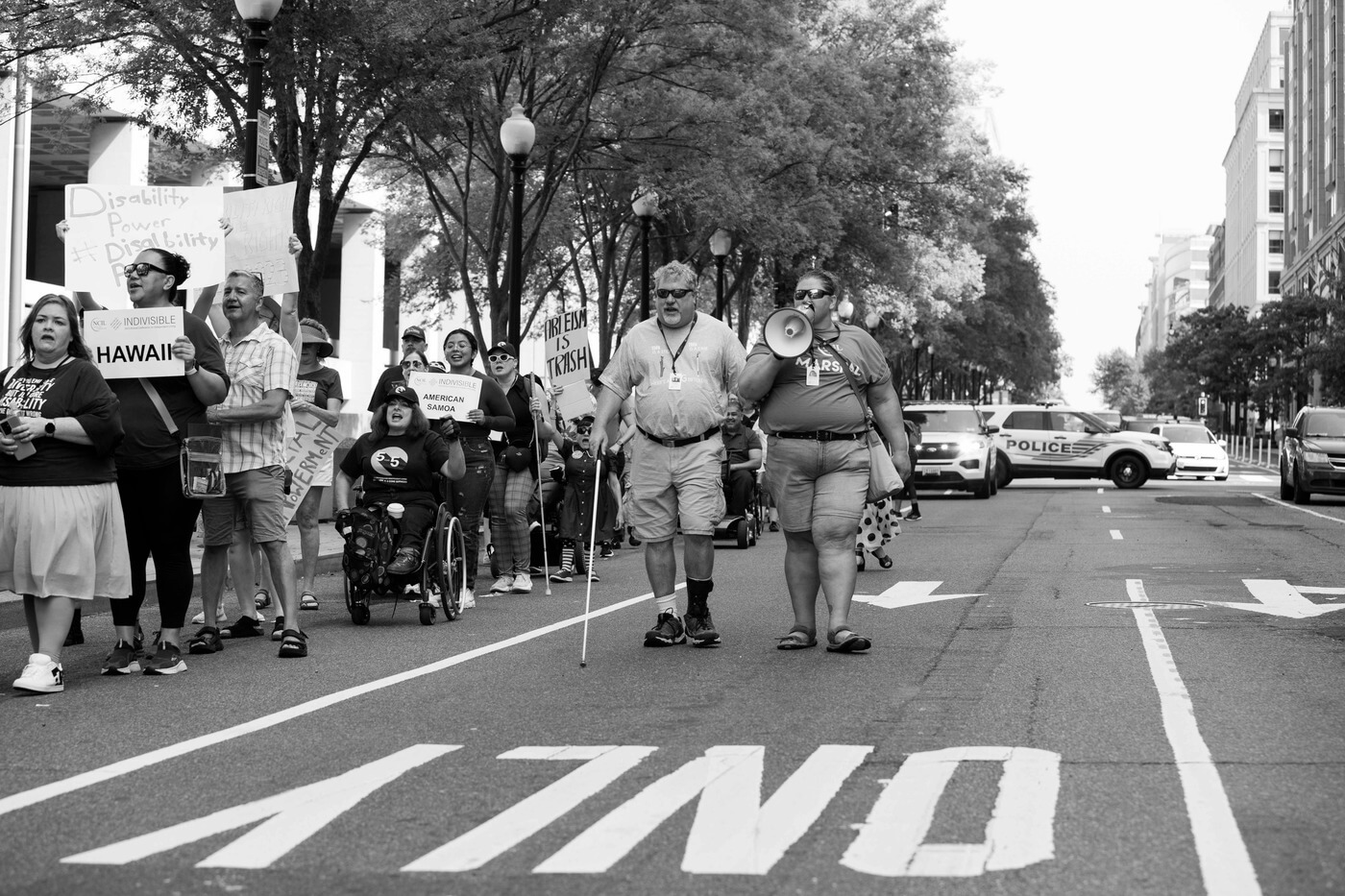 Black and white photo of two advocates walking side by side, one with a long cane and one speaking into a megaphone. Off to the side is a crowd of advocates carrying signs that say things like "Hawaii" "American Samoa" and "Ableism is Trash".