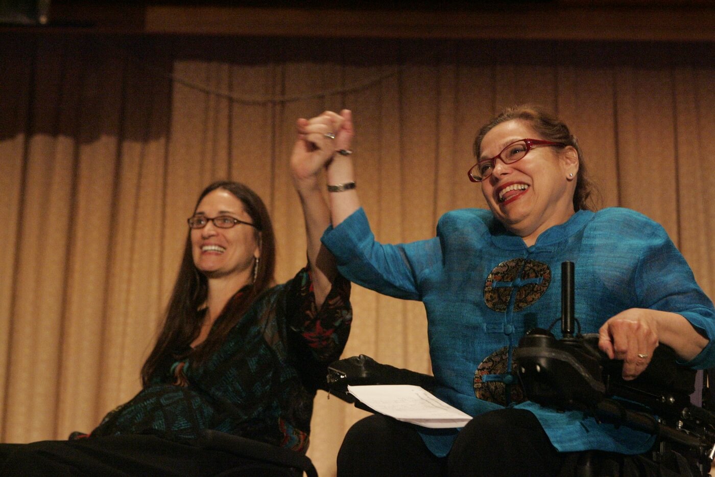 Judy Heumann and Marca Bristo sit on stage together with their hands clasped together and raised.