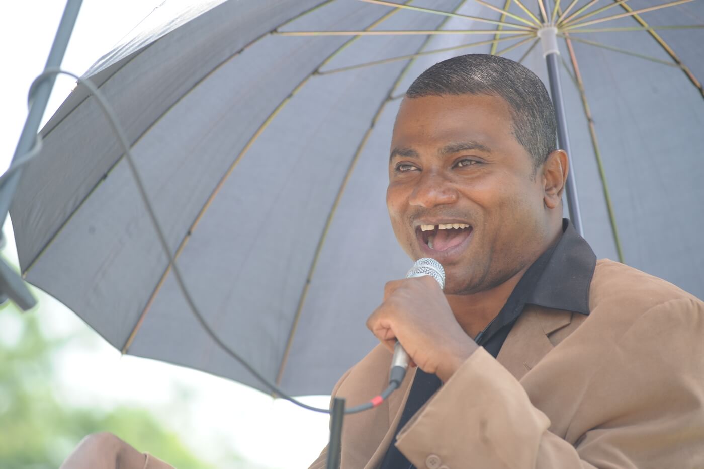 A man with medium brown skin and close cropped black hair stands under an umbrella and speaks into a microphone.