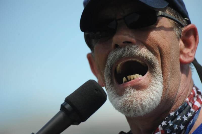 Mark Derry speaks into a microphone at the NCIL Rally. He is wearing a navy cap and an American flag bandana around his neck.