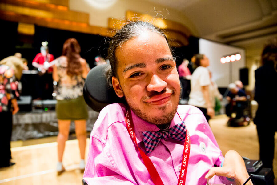 Jensen Caraballo wears a pink collared shirt with pink checkered bow tie and a red lanyard. He is using a power wheelchair and smiles at the camera.