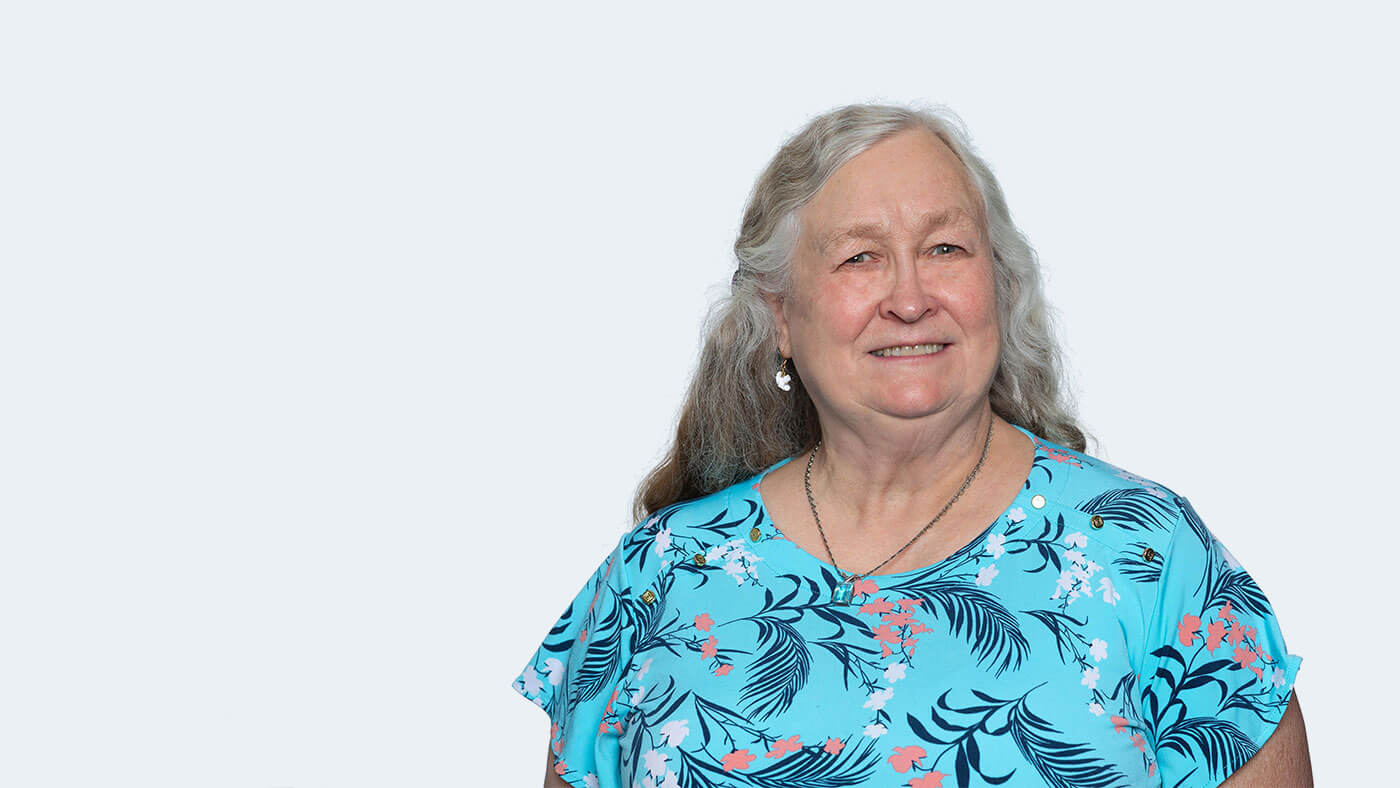 Dee Dee Foremaster, a white person with long wavy grey hair and a floral shirt, smiles at the camera.