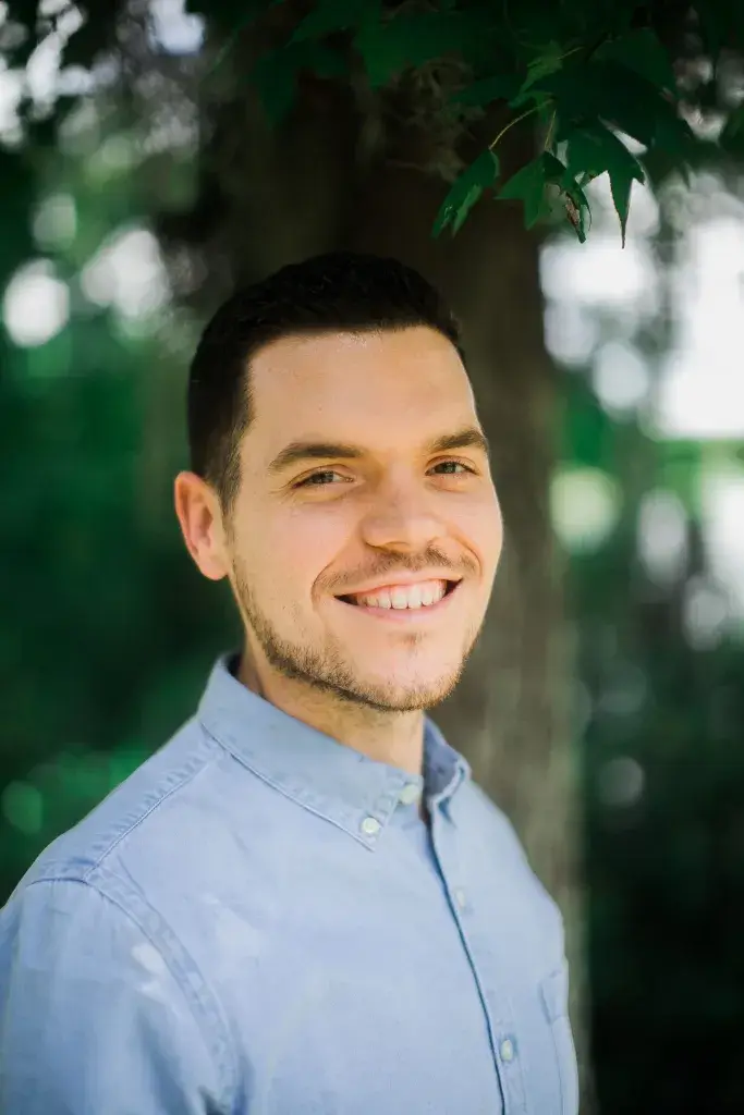 Dom Kelly, a white man with brown hair on his head wearing a light denim collared shirt, standing in front of a tree surrounded by green leaves.