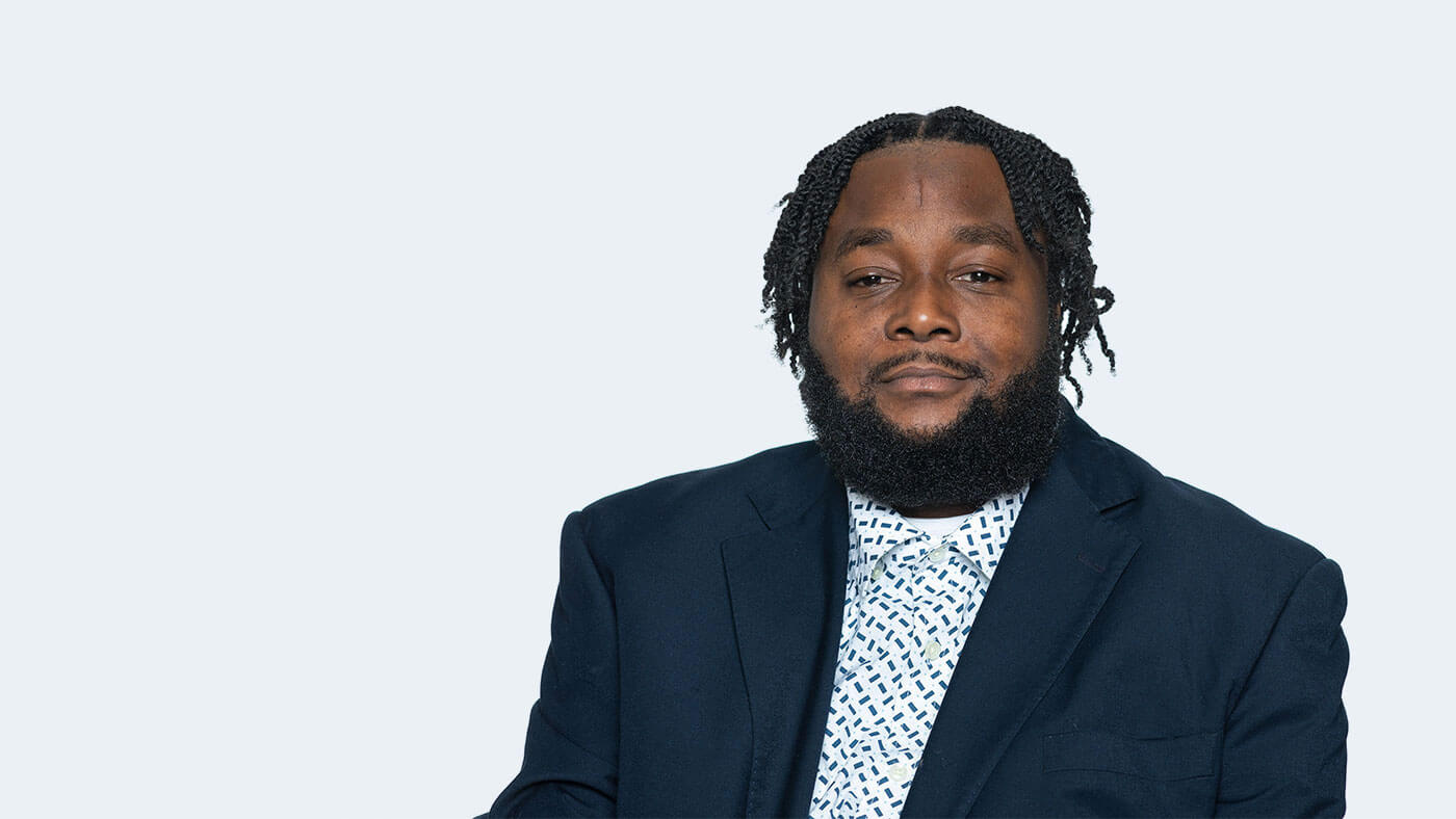 Edward Mitchell, a Black person with short hair in braids and a bushy black beard, looks at the camera.