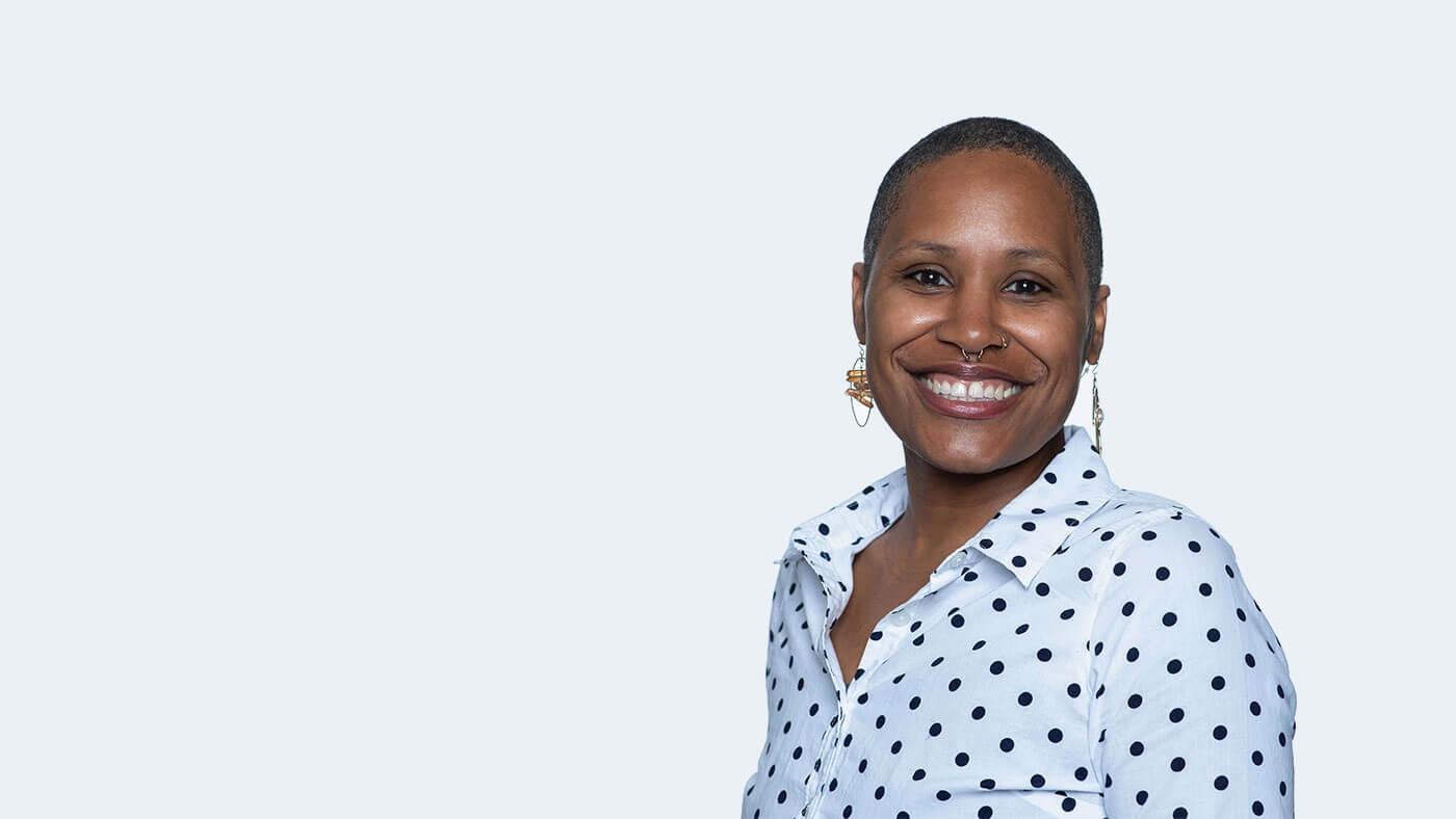 Ester DuRante, a Black person with short, close cropped black hair, a nose ring, and dangly earrings, smiles widely at the camera.