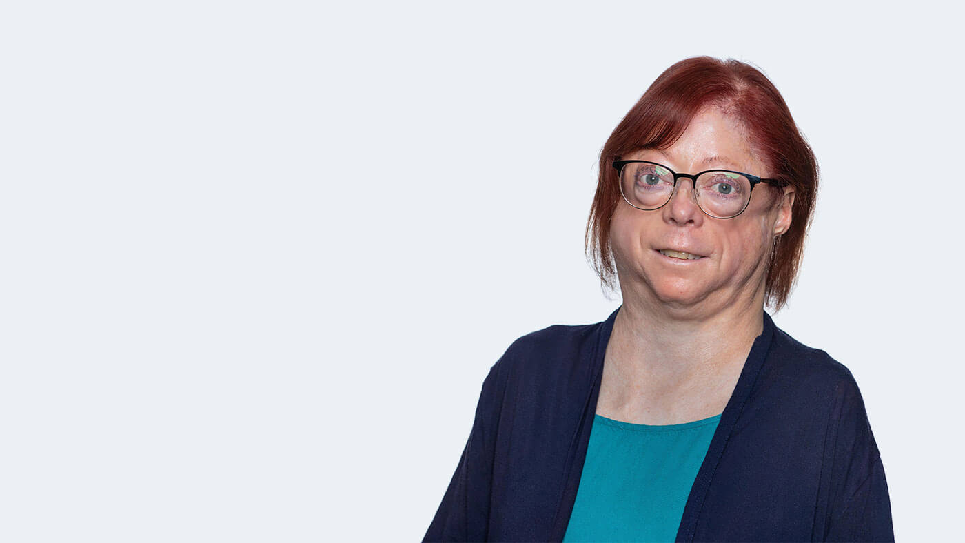 Joan LaBelle, a white person with glasses and short red hair, smiles at the camera.