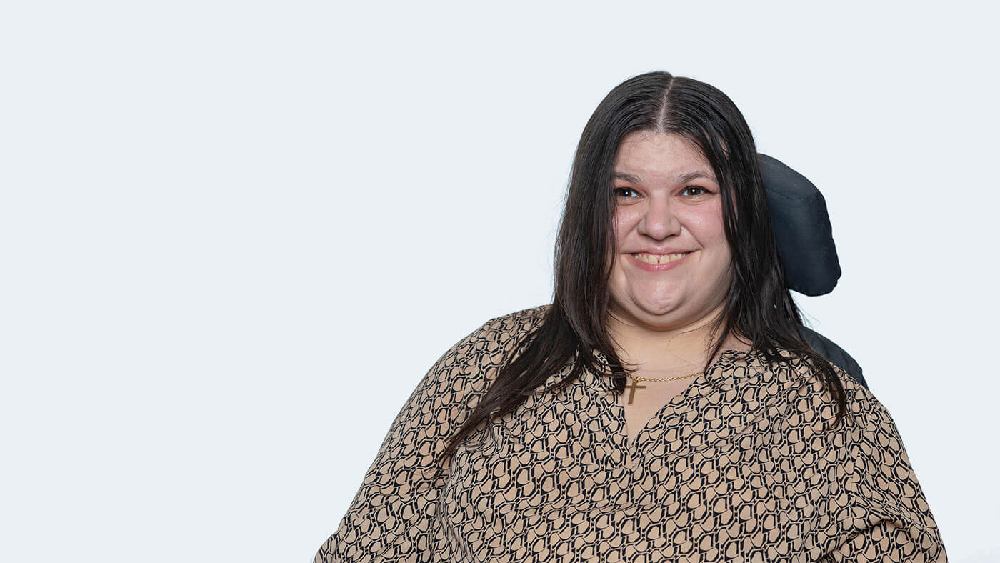 Jordan Hall, a white person with straight black hair just past the shoulders using a wheelchair, smiles at the camera.
