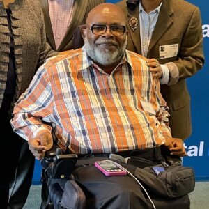 Theo Braddy – A black man with a bald head, a salt and pepper full beard wearing eyeglasses, sitting in a wheelchair. Theo smiles toward the camera during a 2023 speaking event. 
