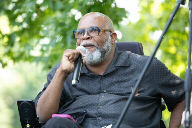 Theo Braddy - A black man with a bald head, a salt and pepper full beard wearing glasses, is speaking into a microphone on stage at NCIL's Rally at the Capitol.