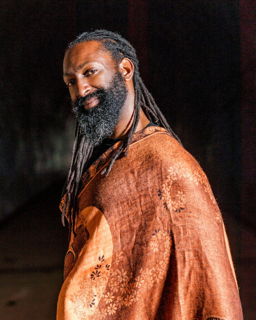 Antoine Hunter: A Native American African-American person with dark chocolate skin from his mother. He has almond shaped eyes with long lashes. He has long black dreadlocks tied in a low ponytail and a full beard. Antoine is wearing a large brown wrap and looking artistically at the camera.