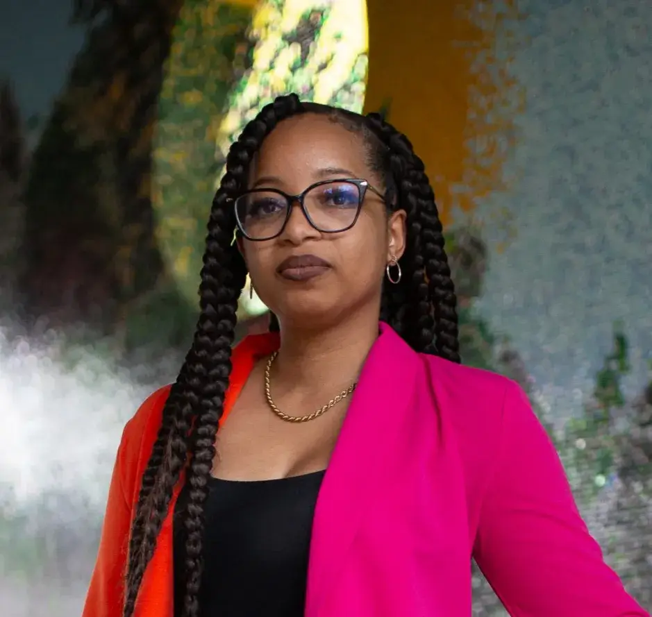 Keri Grey looks boldly into the camera for a headshot with an abstract background. Keri is wearing a neon orange and pink blazer and glasses.
