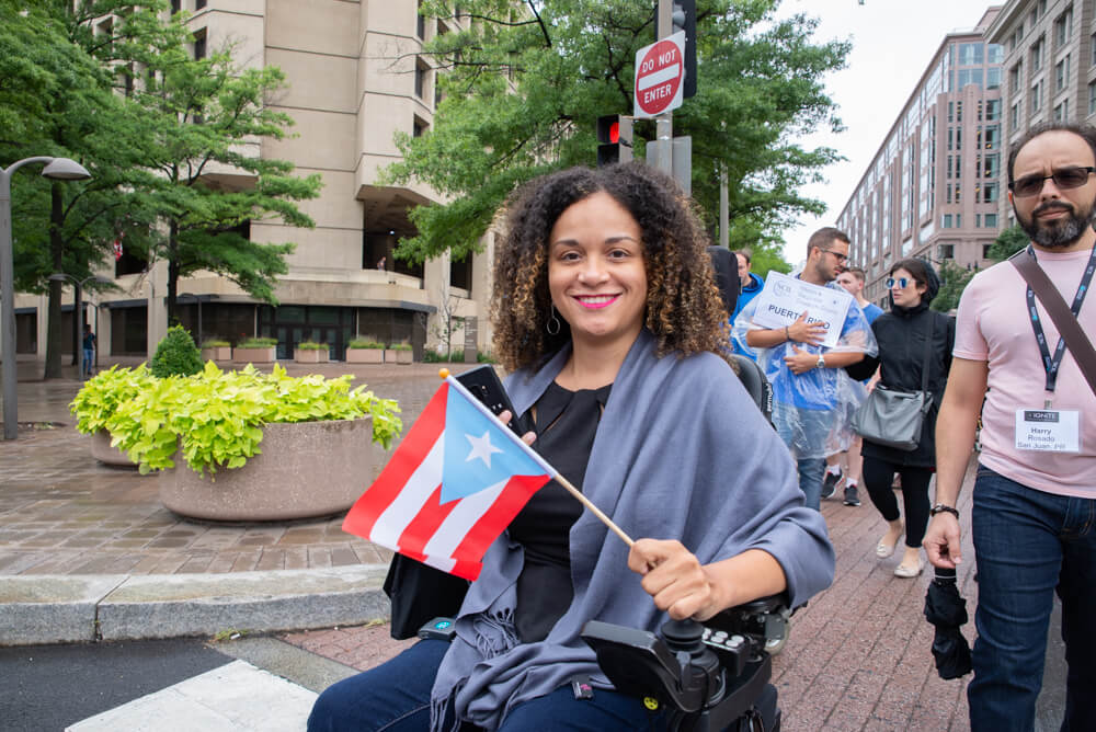 An advocate using a power wheelchair holds a small Puerto Rican flag.