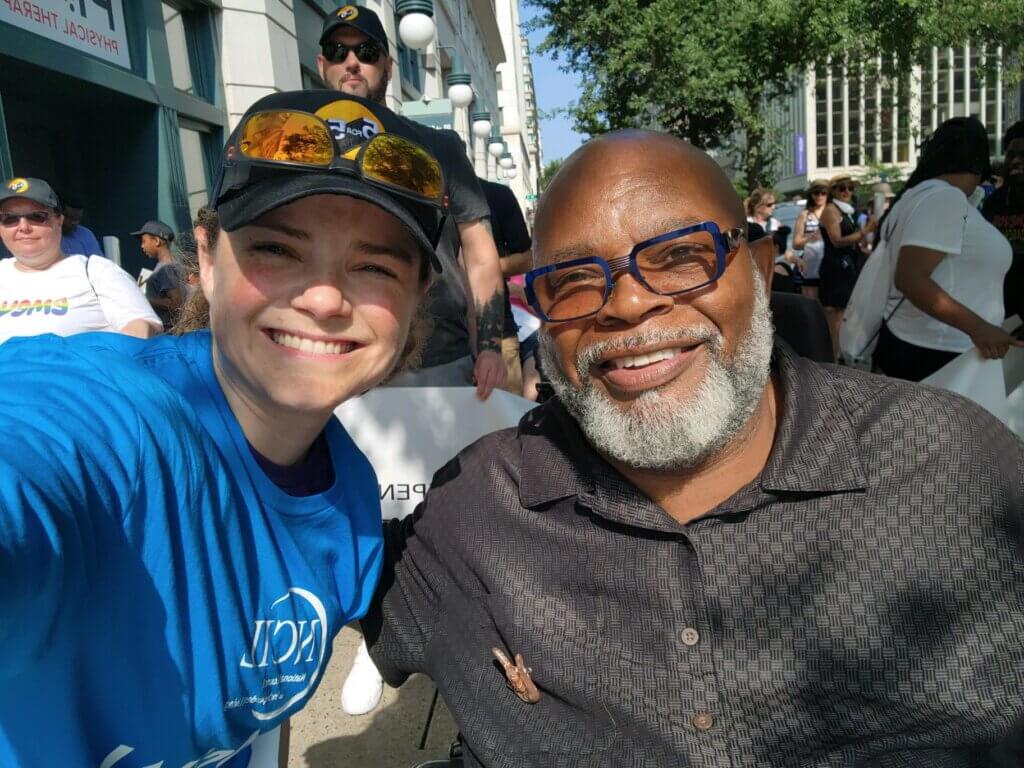 NCIL staff Jenny Sichel and Theo Braddy pose for a selfie outside the Grand Hyatt. Behind them, the sidewalk is filled with people lining up to begin the NCIL 2023 March to the Capitol.