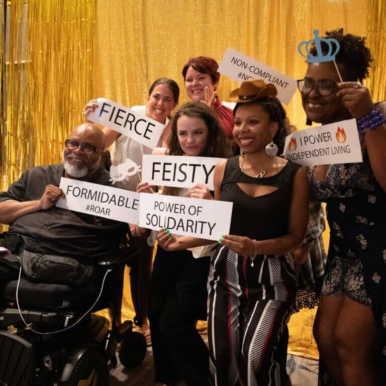 NCIL staff members Theo Braddy, Sylena Campbell, Jenny Sichel, Denise Law, Jessica Podesva, and Mary-Kate Wells pose for a group picture in the photo booth at NCIL’s 2023 Annual Conference on Independent Living. They are holding signs that say Fierce, Feisty, Formidable, Power of Solidarity, Power of Independent Living, and Non-Compliant.