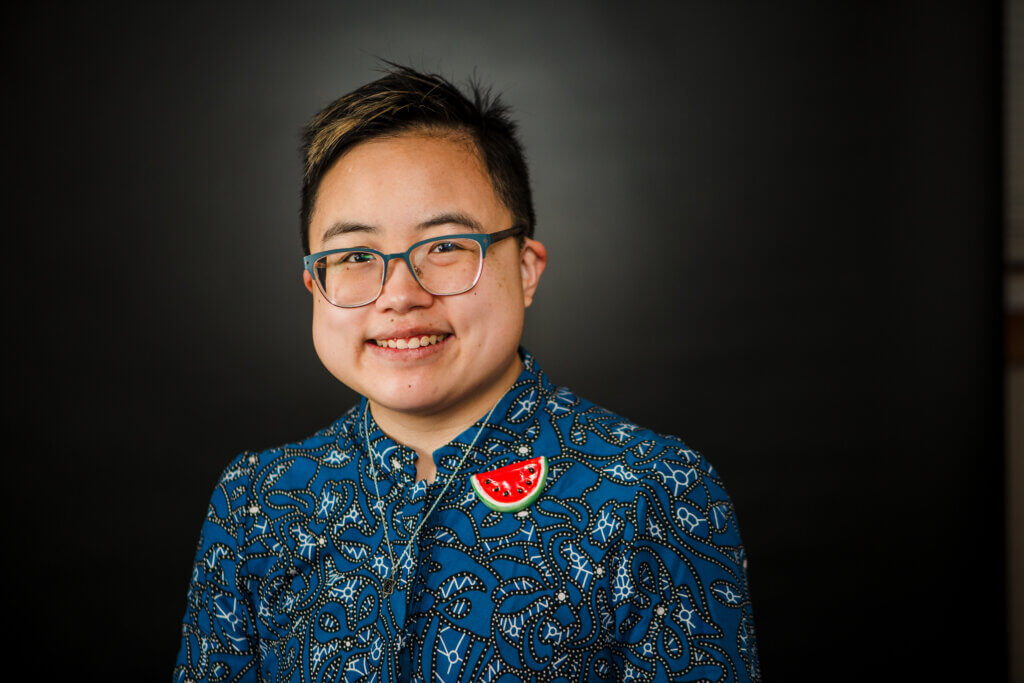 Ly Xīnzhèn is an androgynous and transmasculine East Asian person in their early thirties. They have short black hair with light highlights and they’re wearing a blue top with an abstract pattern, a handmade watermelon pin, and a silver magen david necklace. 