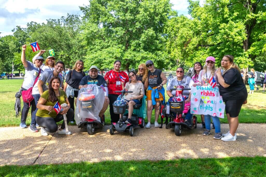 Fourteen advocates gather for a group photo at the 2024 NCIL Rally at the U.S. Capitol. They are holding signs and waving mini flags on the lawn.