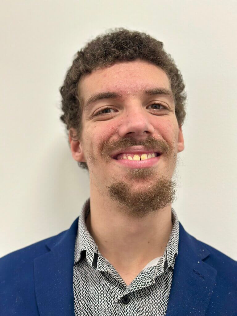 Cody Rooney: Close-up portrait of a light-skinned man in his 20s with short, curly brown hair. He is smiling, and his teeth are visible. He has a short, trimmed brown goatee. He is wearing a navy-blue blazer over a light gray, patterned collared shirt.