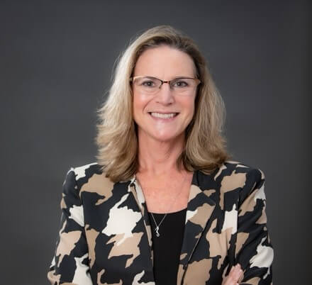 A young woman with medium length light brown hair wearing glasses and a black shirt with a black, tan and cream colored patterned jacket smiling for the camera.