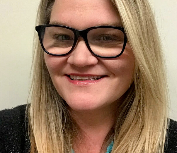 Headshot photo of a white woman with blonde hair and black glasses. She is smiling for the camera.
