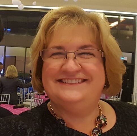 A photo of Isabel Hodge. She is a Caucasian woman and is smiling, has chin-length blonde hair, and wearing costume jewelry.