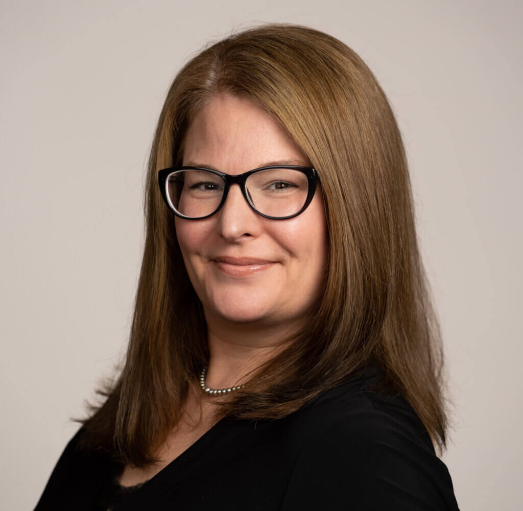 Susan Larsen is a white woman with long, straight brown hair. She wears a black blouse and black-rimmed glasses.